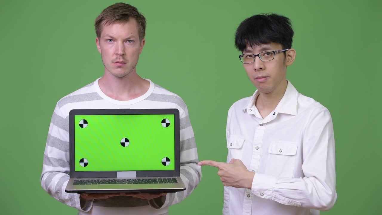 두 명의 젊은 다민족 사업가들이 손가락을 가리키면서 카메라에 노트북을 보여주고 있습니다.