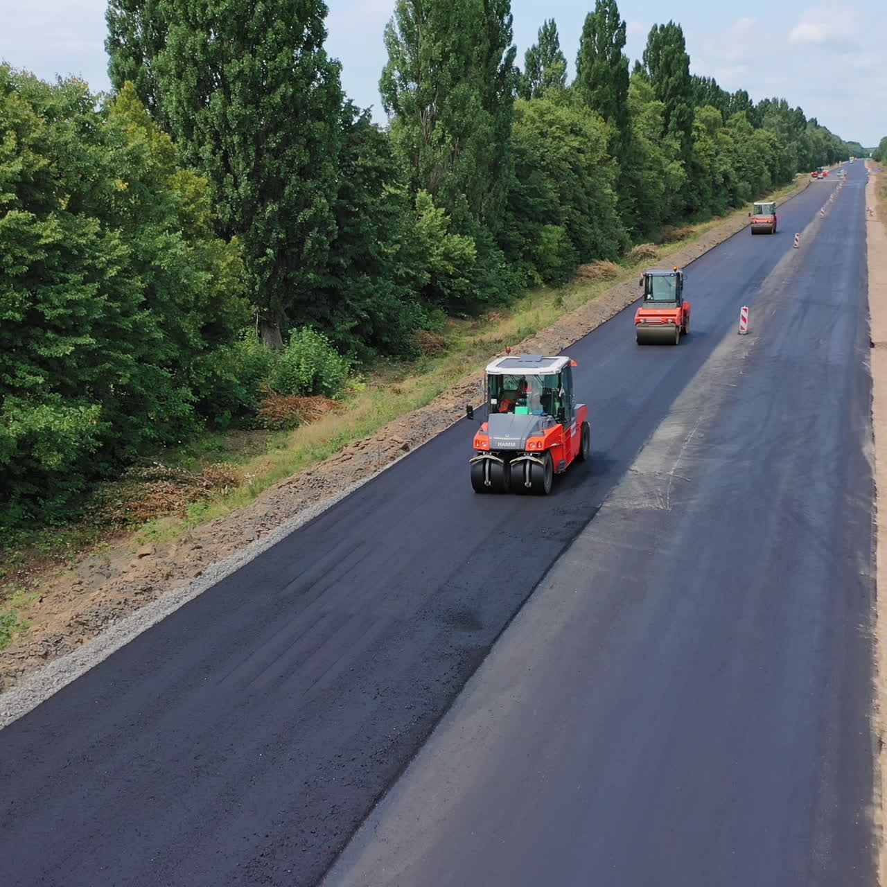 Road rollers working on the highway. Trucks leveling up fresh asphalt on the road among green nature. Asphalt road construction. Aerial view. Motion camera back.