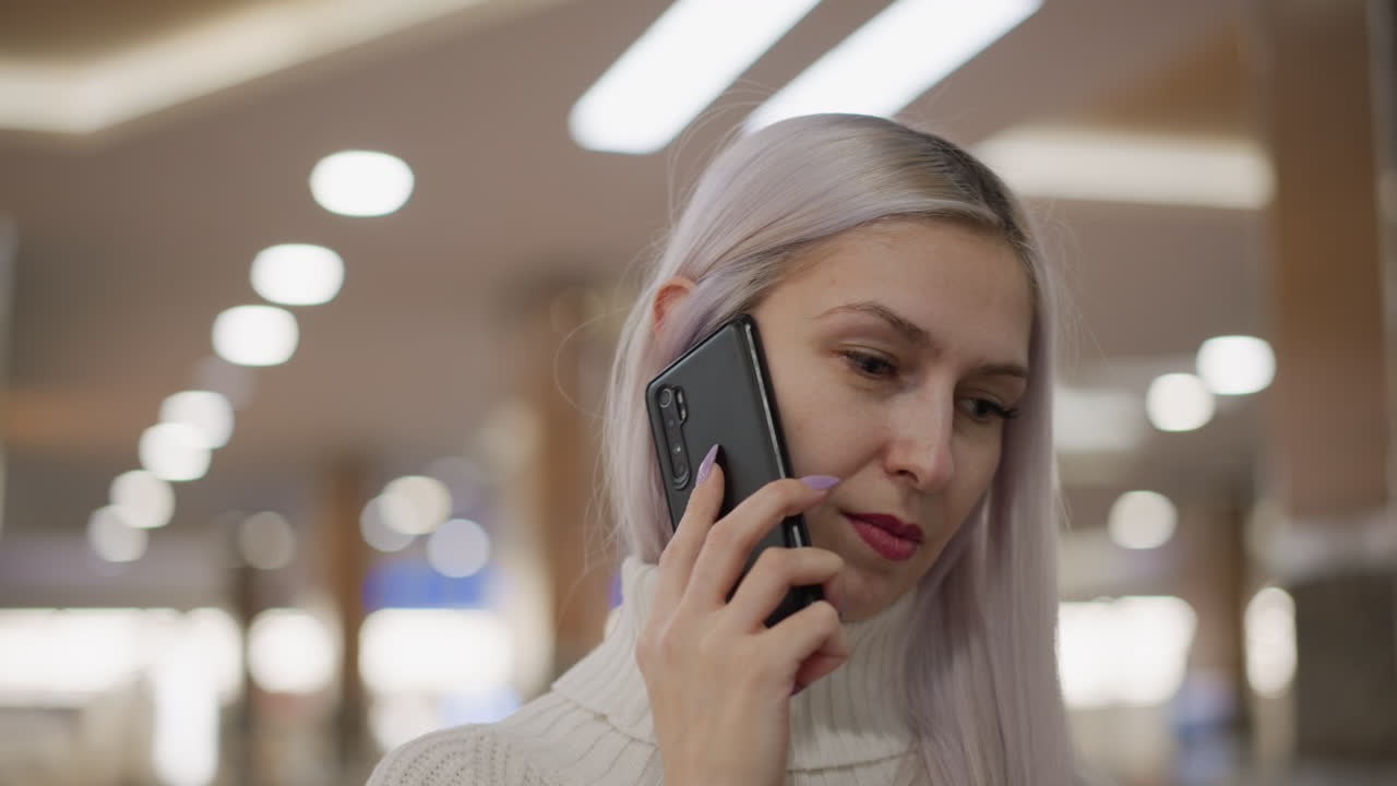 fair skinned woman with long hair standing in mall holding phone to ear while listening thoughtfully against backdrop of blurred lights and glass railing wearing white sweater jeans boots and watch