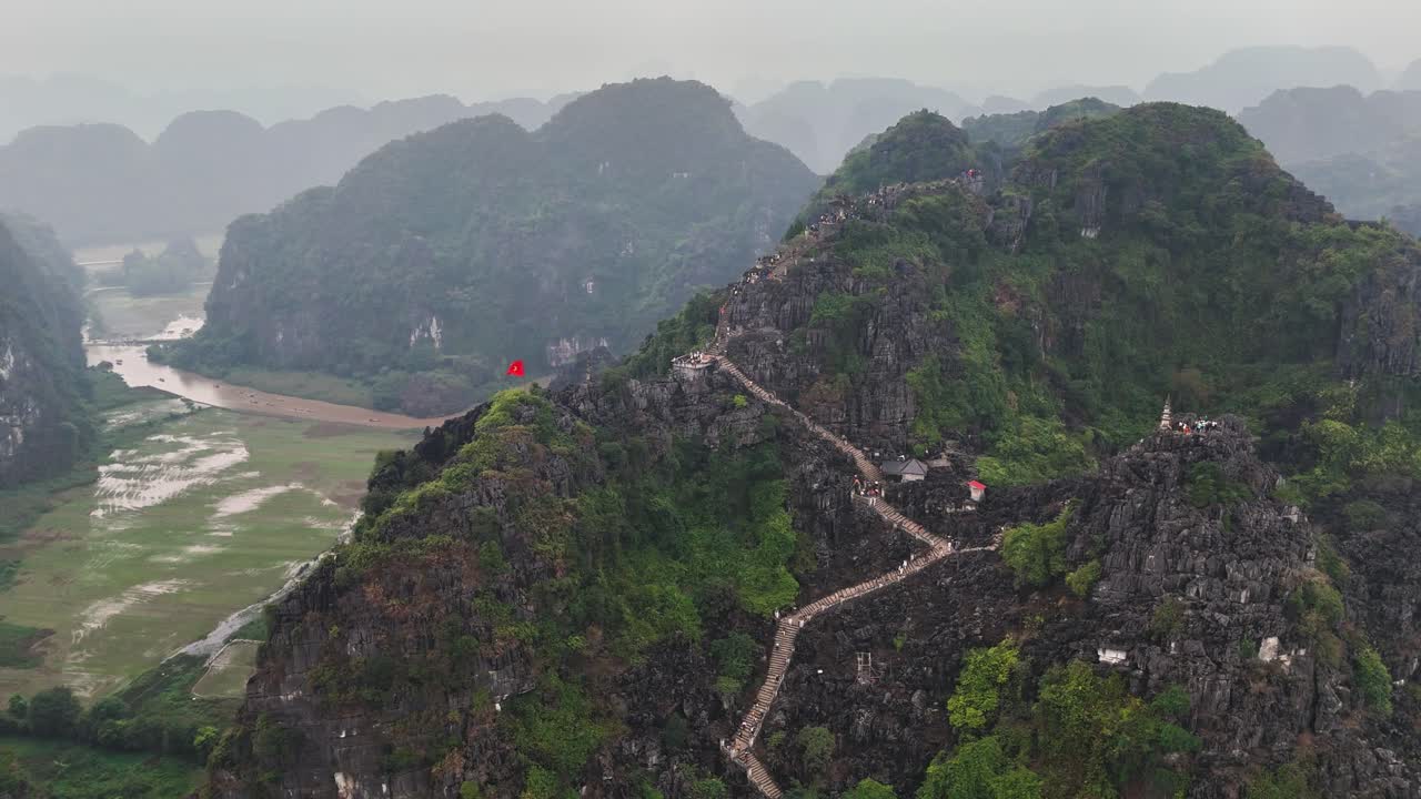 Aerial orbit around Dragon Hill in Tam Coc, Ninh Binh, Vietnam, showing the steep stone staircase, limestone karst peaks, and surrounding rice fields in misty weather