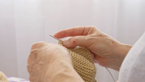 the old woman is holding a knitting needle and a piece of beige knitted clothes. favorite hobby of elderly grandmothers, do-it-yourself knitting at home for their children handmade knitting