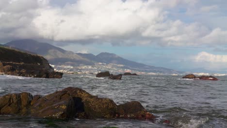 Rocas,-Olas-Del-Mar-Y-Nubes-Con-La-Ciudad-Al-Fondo