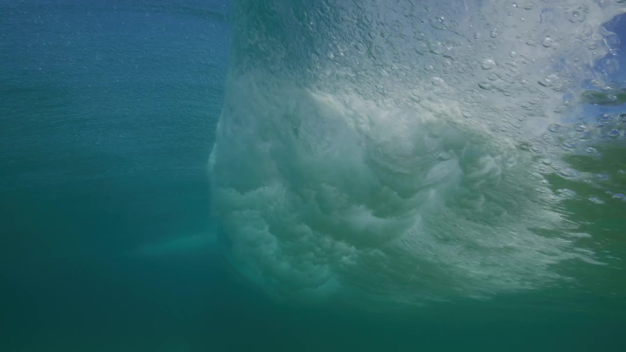 Breaking wave rolls overhead through clear turquoise ocean water with the surface seen from below, rolling vortex throughs spray disrupting calm surface