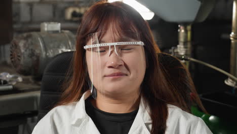 técnico de laboratorio con escudo de protección facial parpadea los ojos con una cálida sonrisa en el taller mecánico, el fondo presenta maquinaria e iluminación brillante, creando un espacio de trabajo profesional