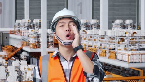 close up of asian male engineer with safety helmet yelling with hand over mouth while standing in the warehouse with shelves full of delivery goods