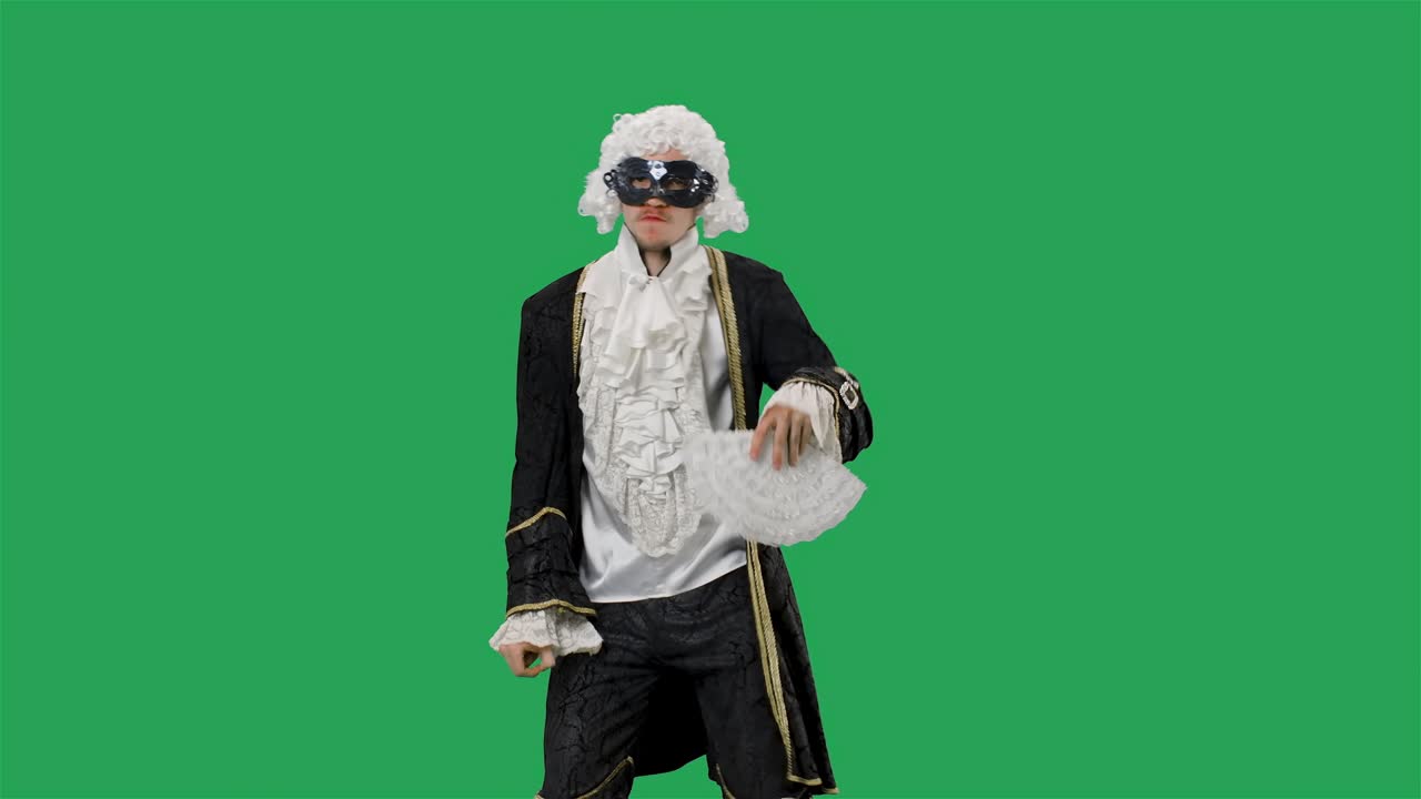 검은색 역사적인 빈티지 슈트, 색 가발과 마스크를 입은 궁정 신사의 초상화, 손에 팬을 들고 즐겁게 춤을 추고, 녹색 스크린 배경으로 스튜디오에서 포즈를 취하는 젊은이.