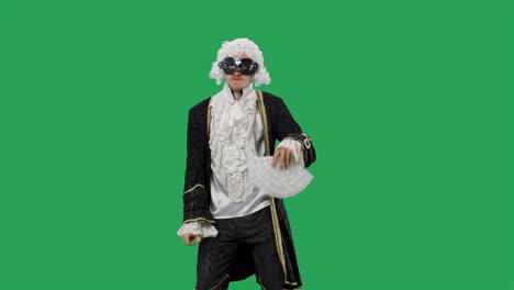 portrait of courtier gentleman in black historical vintage suit, white wig and the mask, dancing merrily with a fan in his hands. young man posing in studio with green screen background. slow motion
