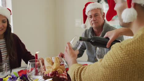 familia caucásica feliz de varias generaciones con sombreros de santa claus, con comida de navidad, vertiendo vino