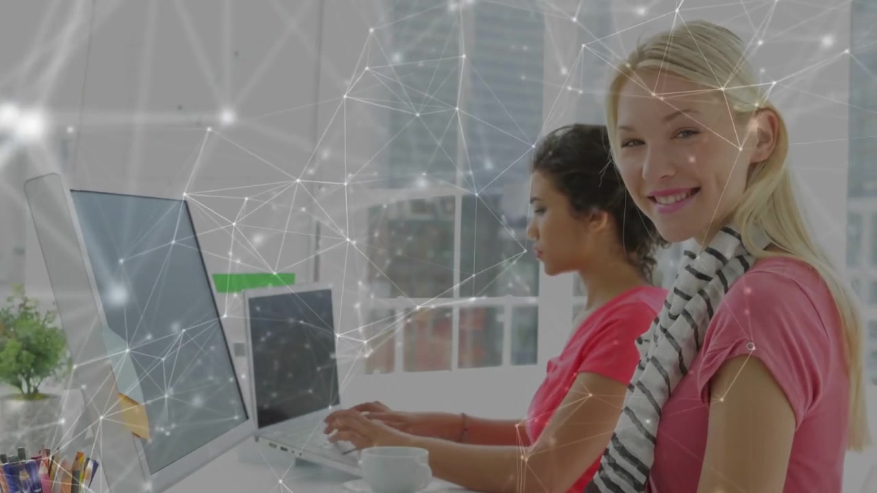 Two coworkers typing at desk, activating network overlay over monitors for business, one smiling