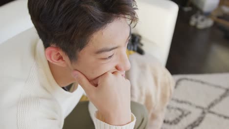 Bored-asian-male-teenager-touching-his-face-and-sitting-in-living-room