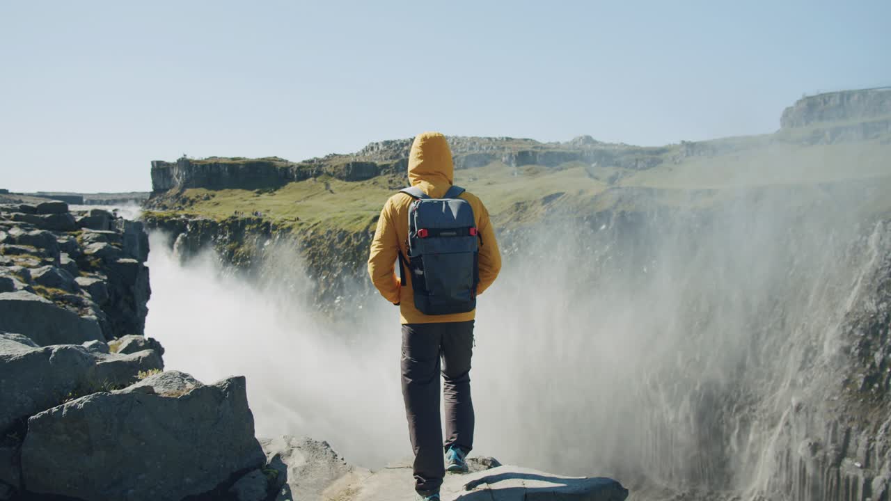 Rear view of man in yellow jacket and backpack standing at cliff edge looking at Detifoss waterfall in Iceland