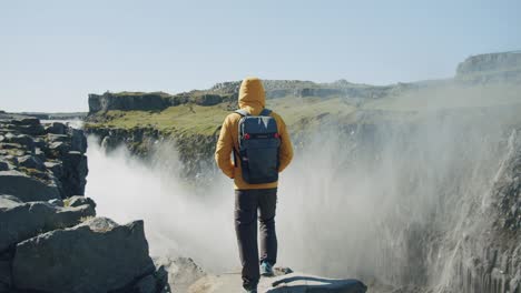 rear view of man in yellow jacket and backpack standing at cliff edge looking at detifoss waterfall in iceland