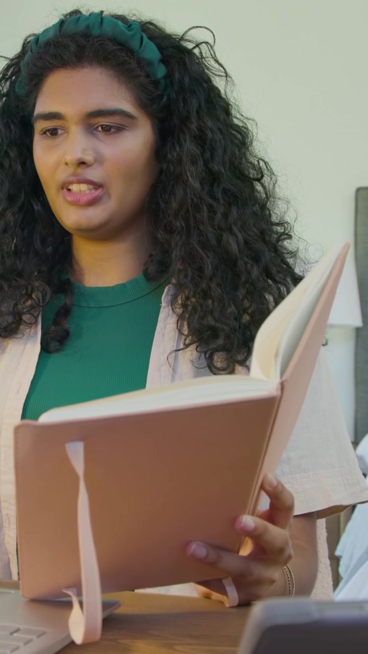 Vertical video: Reading lady in green top showing pink notebook to laptop in bedroom, copy space