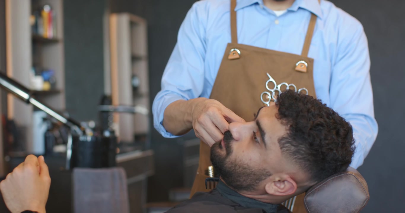 Barbeiro e cliente masculino reclinados, barbeiro aparando o bigode com aparador na barbearia.