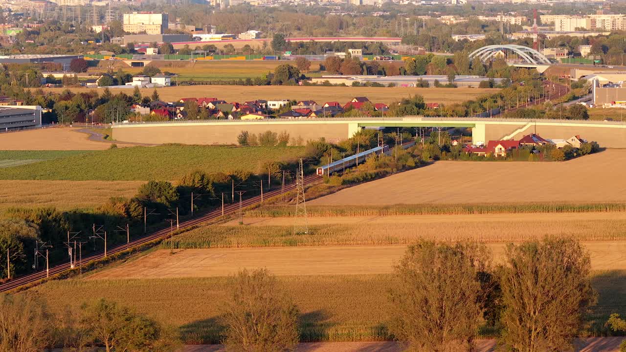 Train on railway between colored farm fields in suburb of Warsaw city during golden sunset. Aerial wide shot. Polish agricultural landscape in rural area. Riding train in the evening
