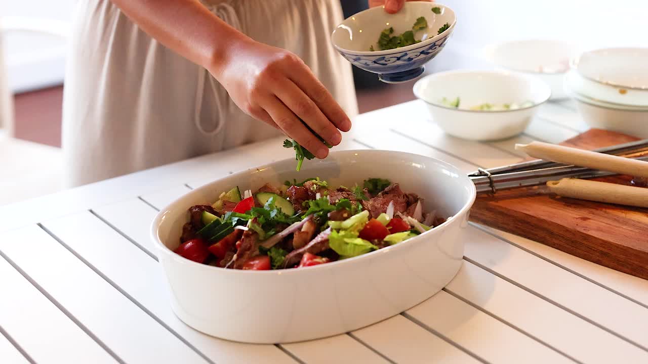 Hands adding coriander to a colorful beef salad in a bright kitchen setting, emphasizing freshness and culinary artistry