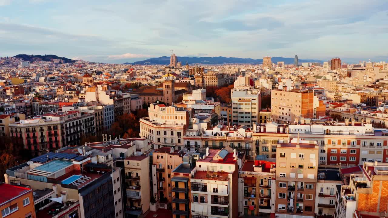 ゴールデンアワーのスペイン、バルセロナの空撮都市景観。密集した屋根が広がり、スカイラインにはサグラダ・ファミリアとトーレ・グロリアスがそびえ立ち、その背景には雄大な山々が連なります。