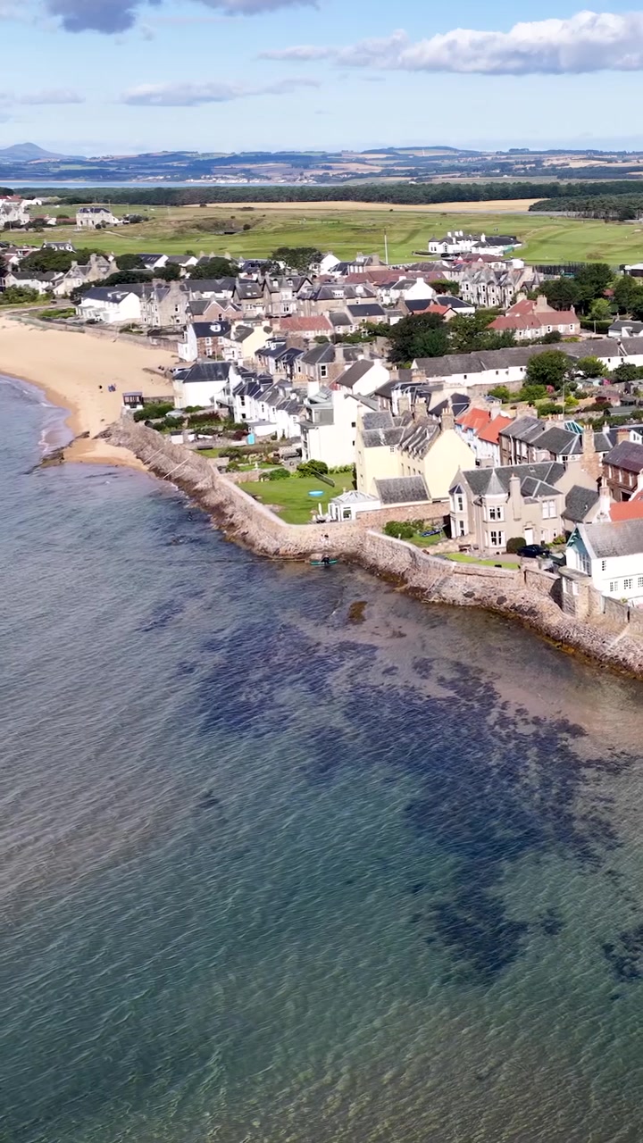 Drone svæver over kystlandsby, sandstrand og huse ved vandet under en lys daghimmel