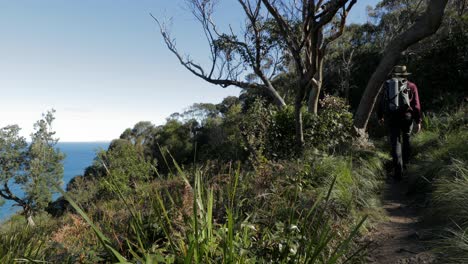 Man-in-hat-walking-away-on-cliff-top-path,-with-ocean-below-to-the-left-hand-side