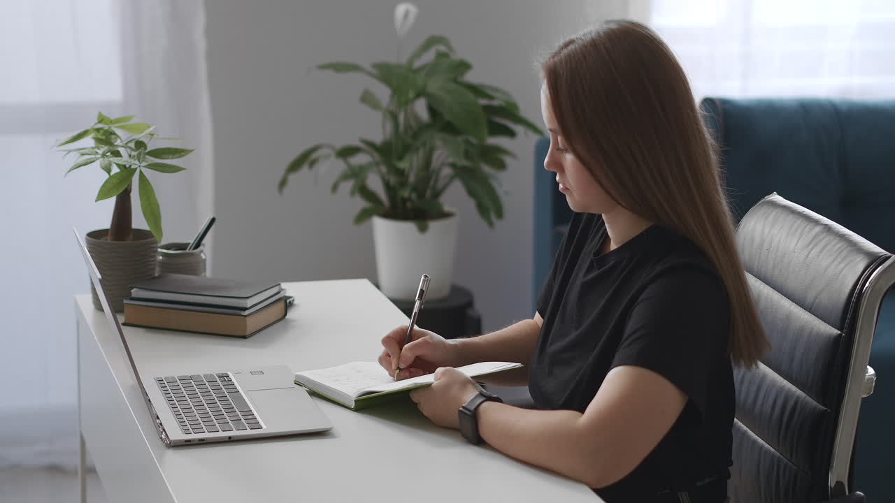 mujer está viendo conferencias en línea escribiendo notas en un cuaderno aprendiendo desde casa educación a distancia y desarrollo profesional sentada en una mesa con una computadora portátil en el apartamento