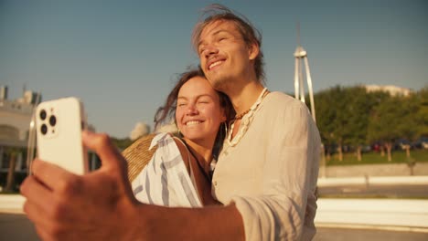 Ein-Glückliches-Paar,-Ein-Mann-Und-Eine-Frau-Machen-Ein-Selfie-Mit-Einem-Weißen-Telefon-An-Einem-Modernen-Strand.-Ein-Glückliches-Date,-Ein-Mann-In-Einem-Weißen-Hemd-Und-Ein-Braunhaariges-Mädchen-In-Einem-Weiß-blauen-Hemd-Machen-Ein-Selfie-Mit-Einem-Weißen-Telefon-An-Einem-Modernen-Strand.