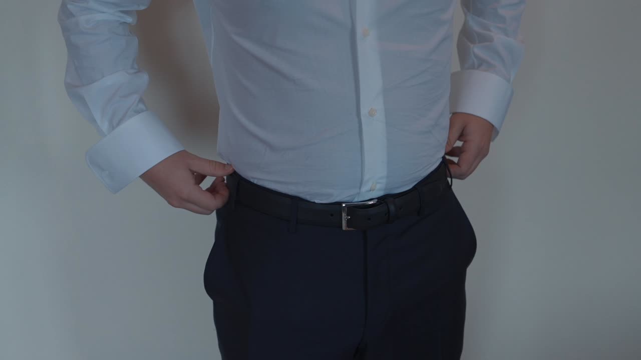Groom adjusting black belt over white shirt and dark formal trousers