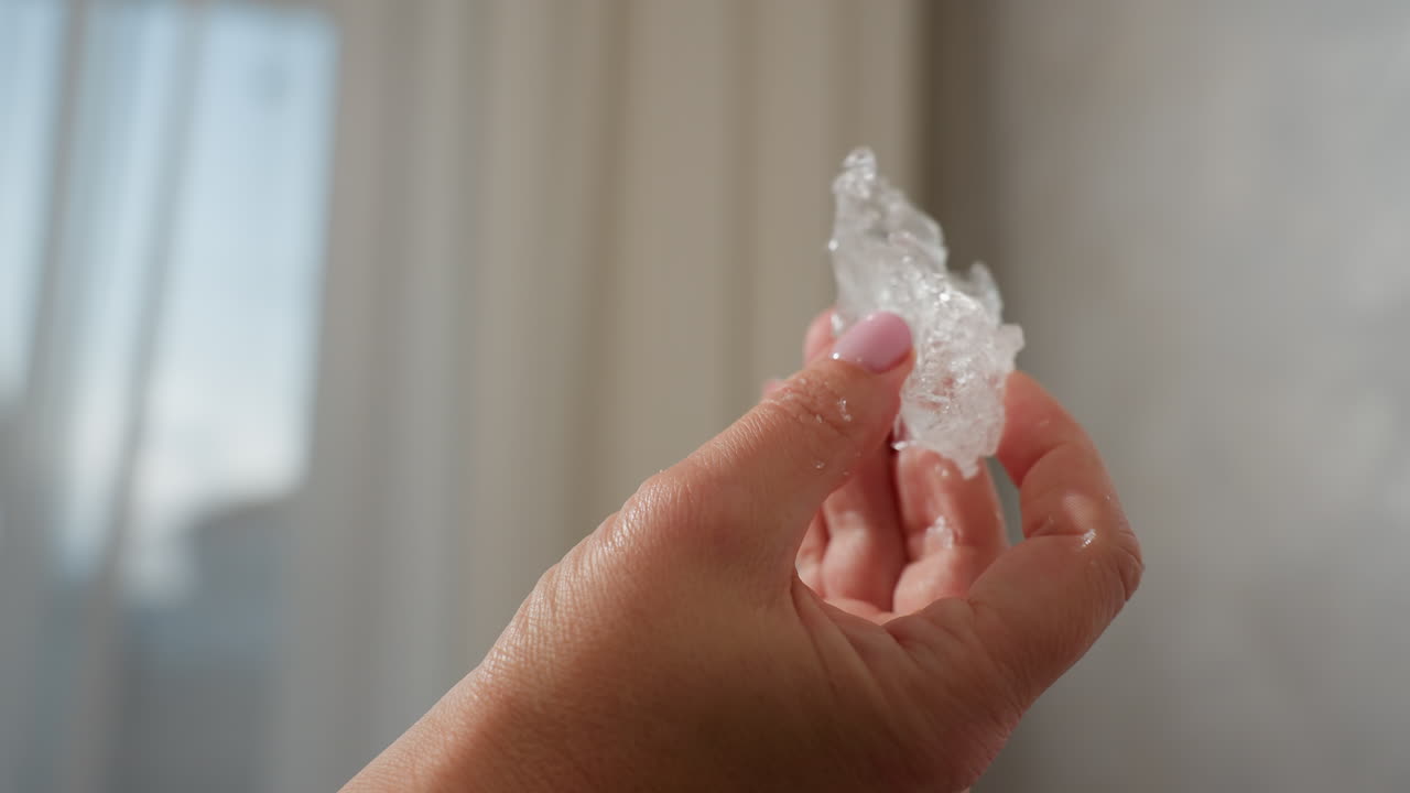 Close up of hand holding clear rubber-like soluble material with water droplets, pink polished nails, natural daylight shining on translucent texture, against soft blurred indoor background