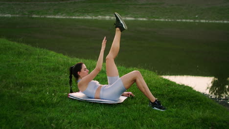 in the morning in the park a woman lying on a mat raises her arm and leg training the abdominal muscles against the background of the river