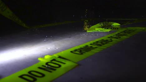slow motion footage of raindrops falling on an abandoned yellow crime scene tape that is on a water covered concrete ground during night time lit by a flashlight. the water is splashing and flying