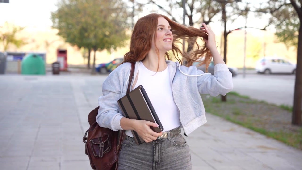 Una giovane donna cammina con sicurezza in un ambiente urbano portando un quaderno e uno zaino.