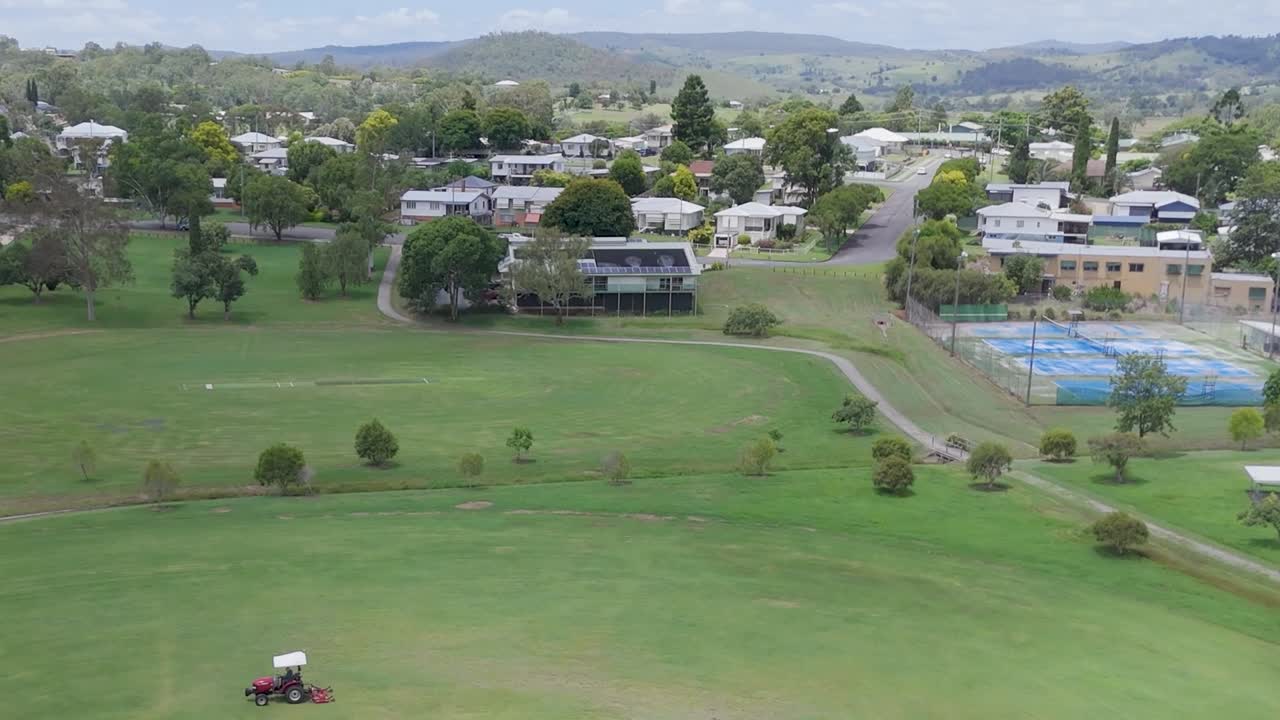 Aerial view of a tractor mowing a large green field adjacent to a neighborhood with visible houses and trees.