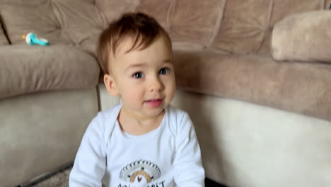 cute dark-haired baby boy in white clothes plays on the floor with soft pillow. kid stands up standing at the sofa.