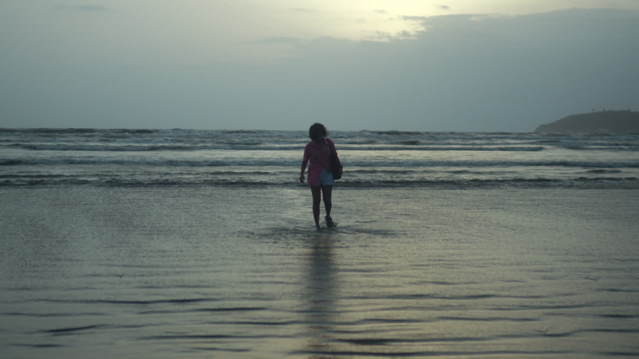 Ampia inquadratura di una donna che cammina da sola nell'acqua bassa del mare sotto un cielo nuvoloso, con onde leggere e una costa distante sullo sfondo.