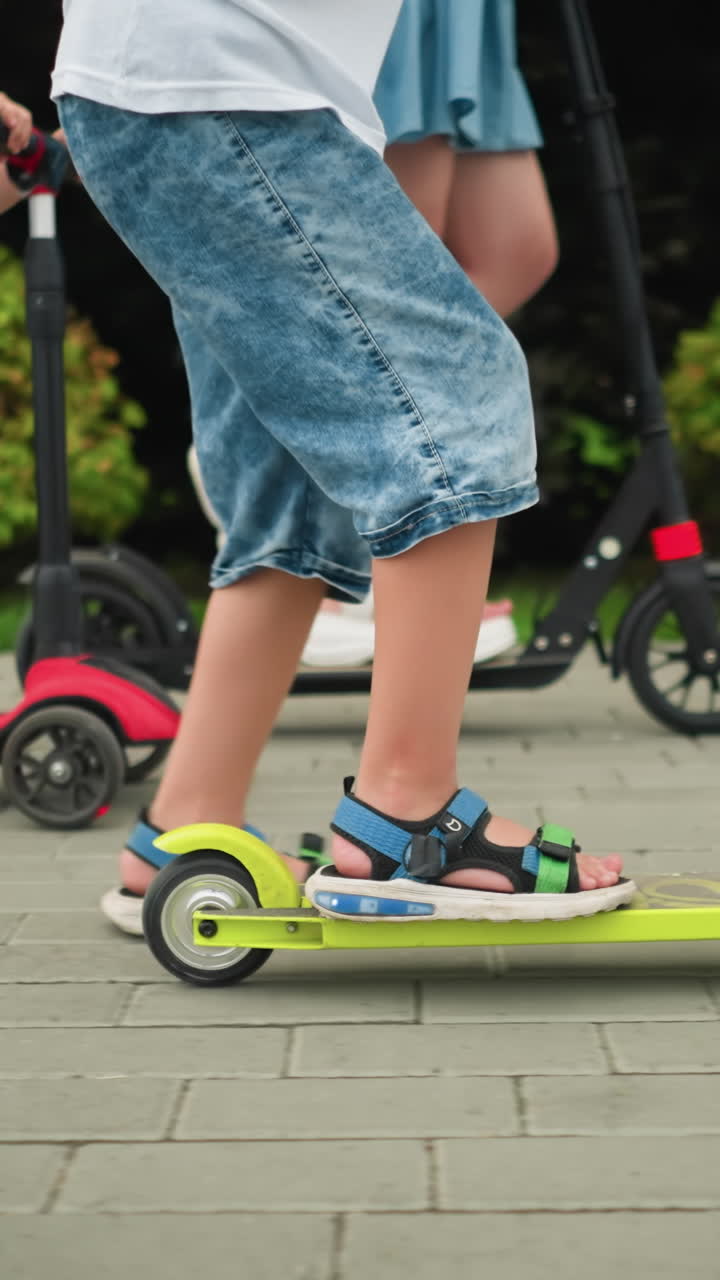 vista de la pierna de una mujer y las piernas de sus hijos mientras viajan en scooters, cada uno usando sus piernas para propulsarse hacia adelante, el niño más joven trabaja duro para alcanzar al resto
