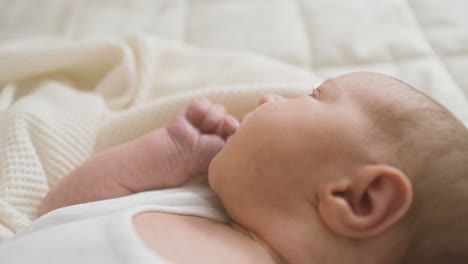 close up view of a baby in white bodysuit lying on bed thumbsucking and moving his arms 1