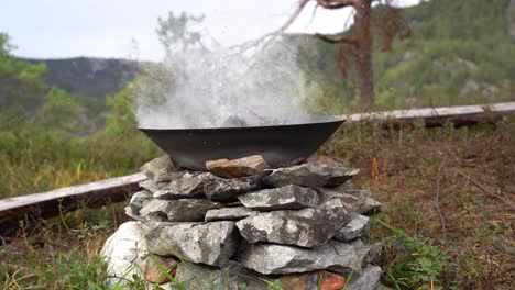 campfire safely extinguished by water to prevent forest fire - slowly pouring water from kettle on top of bonfire inside metal pan - smoke and steam rising up - static nature clip