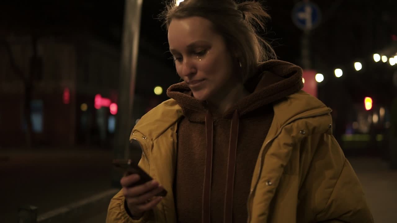 Short haired girl walks through the evening city smiling, stops and texting in the smartphone. Handheld footage