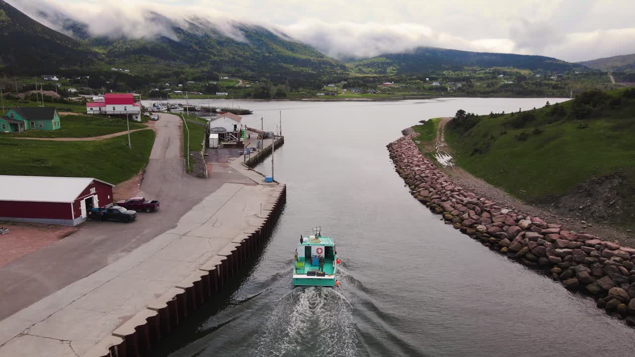 Drone shot following a small commercial lobster fishing boat coming in after a long day revealing a beautiful small village with mountains in the background on Cape Breton Island, Nova Scotia