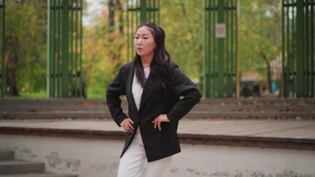 Primo piano di una giovane donna in abbigliamento professionale che posa con sicurezza con le mani sui fianchi all'aperto. La donna indossa un blazer nero e pantaloni bianchi, in piedi contro uno sfondo di parco autunnale.
