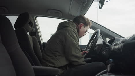 side view of young man in winter jacket adjusting seat while holding steering wheel inside parked car with blurred snowy background visible through window during cold season