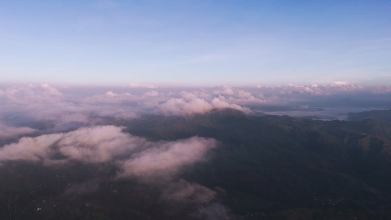 High-angle aerial drone shot capturing a rugged, lush mountain valley and deep canyons, featuring winding roads, small remote villages, and clouds of mist during the dramatic light of golden hour