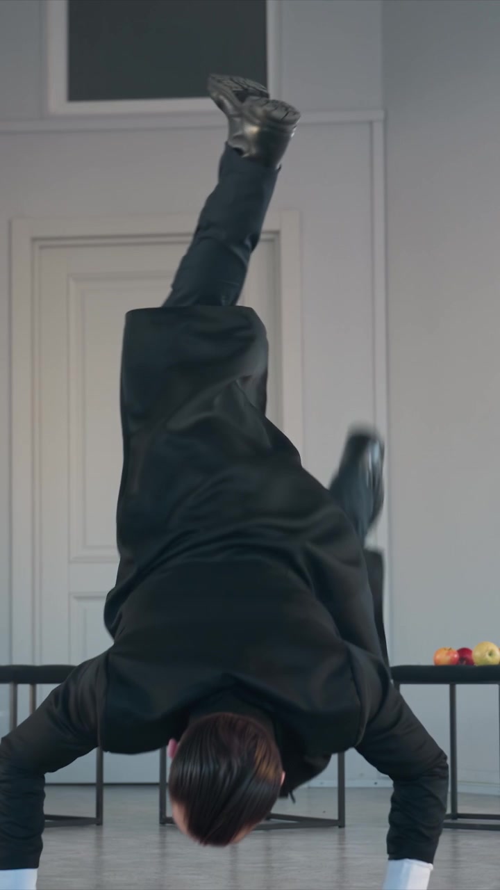 Ballerino esegue un movimento fluido contro una parete chiara. La mano destra si solleva all'altezza del petto mentre il soggetto guarda verso l'alto. Due piccoli tavoli affiancano il soggetto, contenenti mele rosse e gialle.