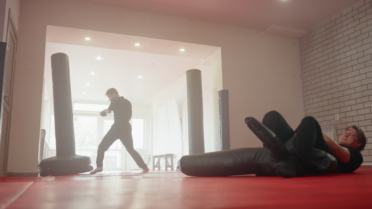 Policeman performing ground combat training exercise on dummy inside gym while another man stands in background observing session, showcasing intensity, martial arts discipline, focus, tactical preparation