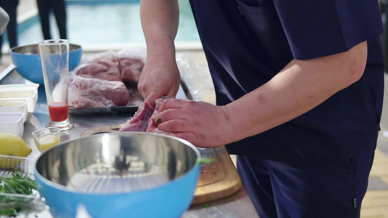 Person's hands cutting a large piece of raw meat using a butcher's knife on a wooden surface outside. Barbeque preparation. shot