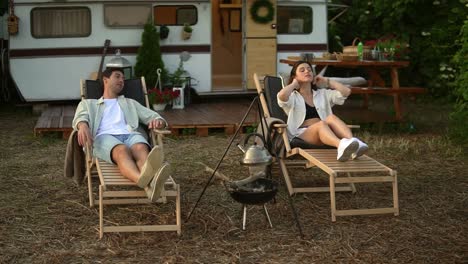 happy young couple having rest close to a campsite with hanged kettle . picnic of a beautiful couple in love at the trailer, laying comfortably on trestle beds. rest in nature. modern trailer on background