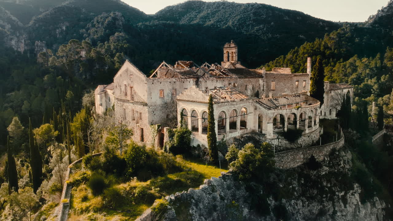 Ripresa cinematografica aerea 4K con drone, in movimento verso il sole, sopra un monastero abbandonato in Catalogna, Spagna. Complesso storico in pietra circondato da montagne spettacolari e foreste all'ora d'oro.