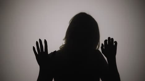silhouette of woman emerges from smoke and touches glass with her palms - silhouette of woman emerges from smoke and touches glass with her palms. woman examines glass and slides her hands over it with interest. model poses in dark studio with yellow soft lights in background. close up