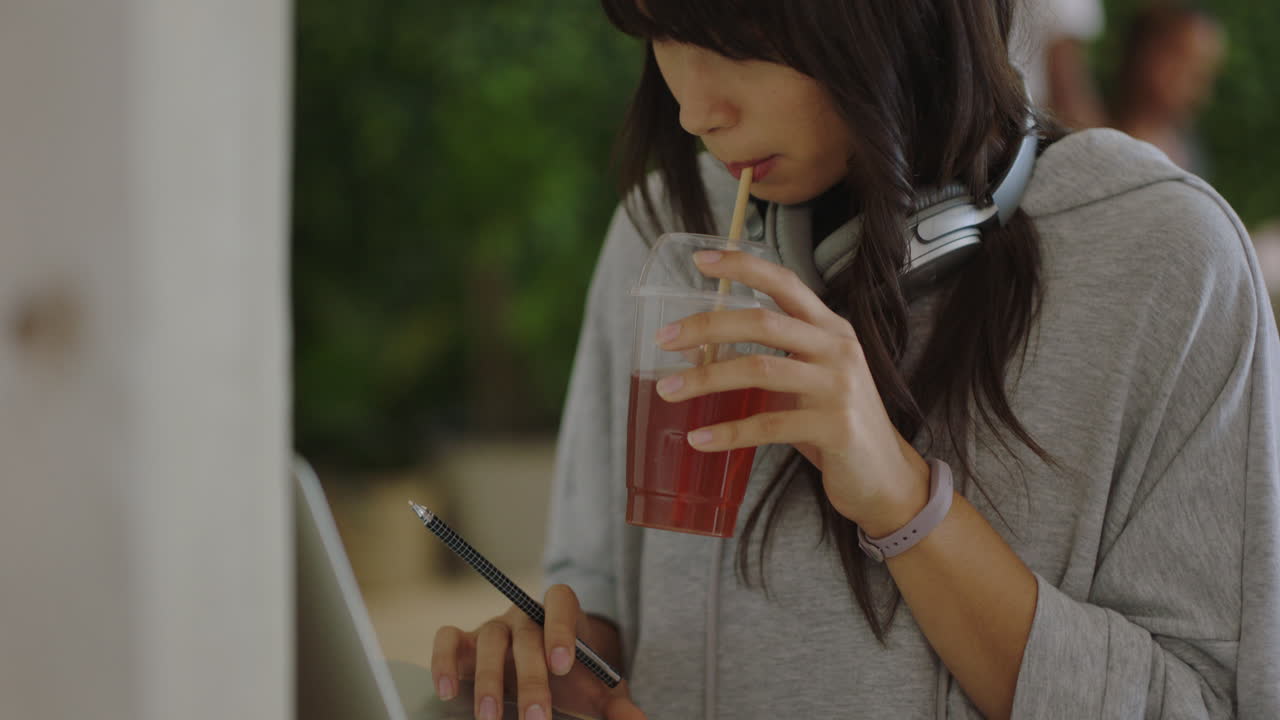 joven mujer de negocios asiática usando una computadora portátil trabajando en un proyecto bebiendo jugo disfrutando de un proyecto creativo en un ocupado lugar de trabajo de oficina
