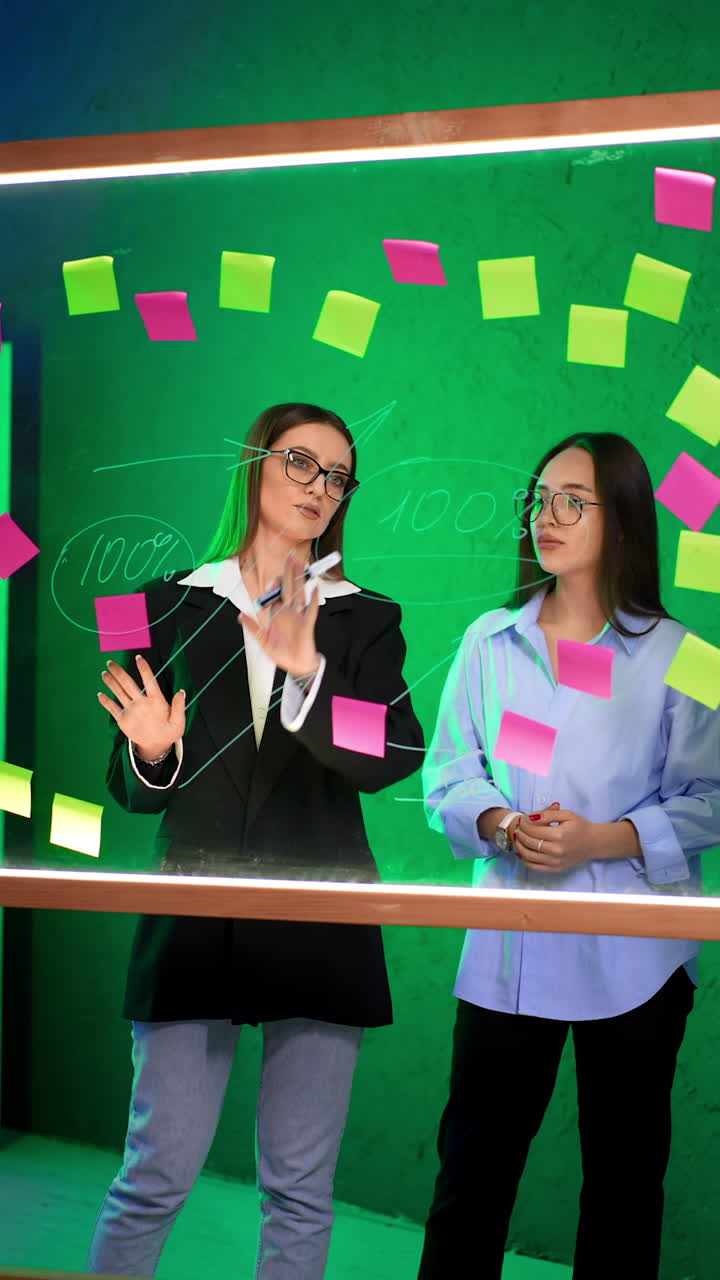 Caucasian women communicate standing behind the glass board. Ladies discuss numbers on the glass wall planner in boardroom, brainstorming. Vertical video.