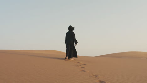 back view of an unrecognizable muslim woman with hijab walking barefoot in a windy desert